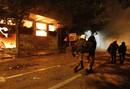 Agentes antidisturbios acompa&ntilde;an a dos detenidos mientras pasan por un edificio hist&oacute;rico de Atenas en llamas